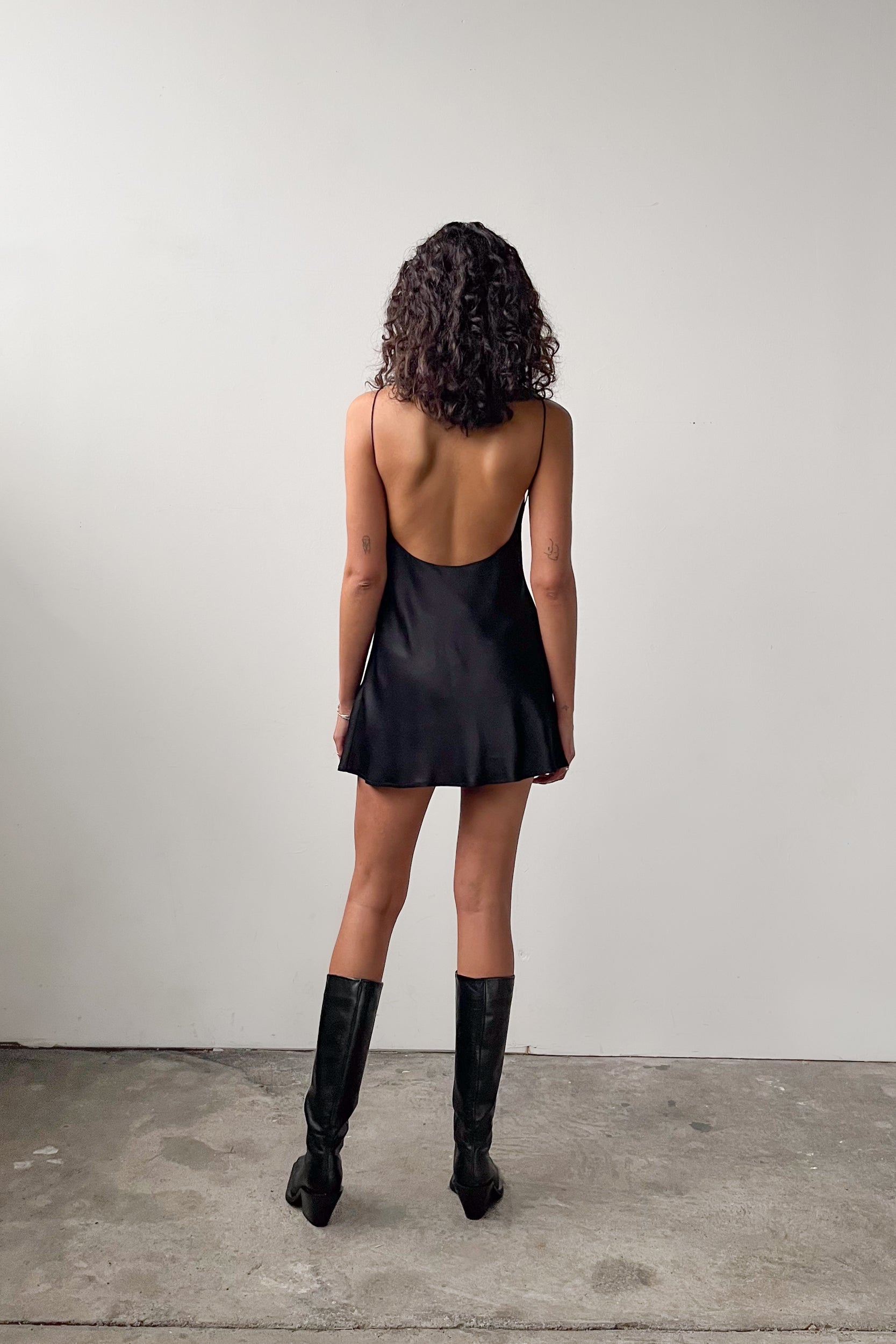 Back view of the Backless Short Slip Dress in Black, showcasing its open back design and fluid satin fabric.