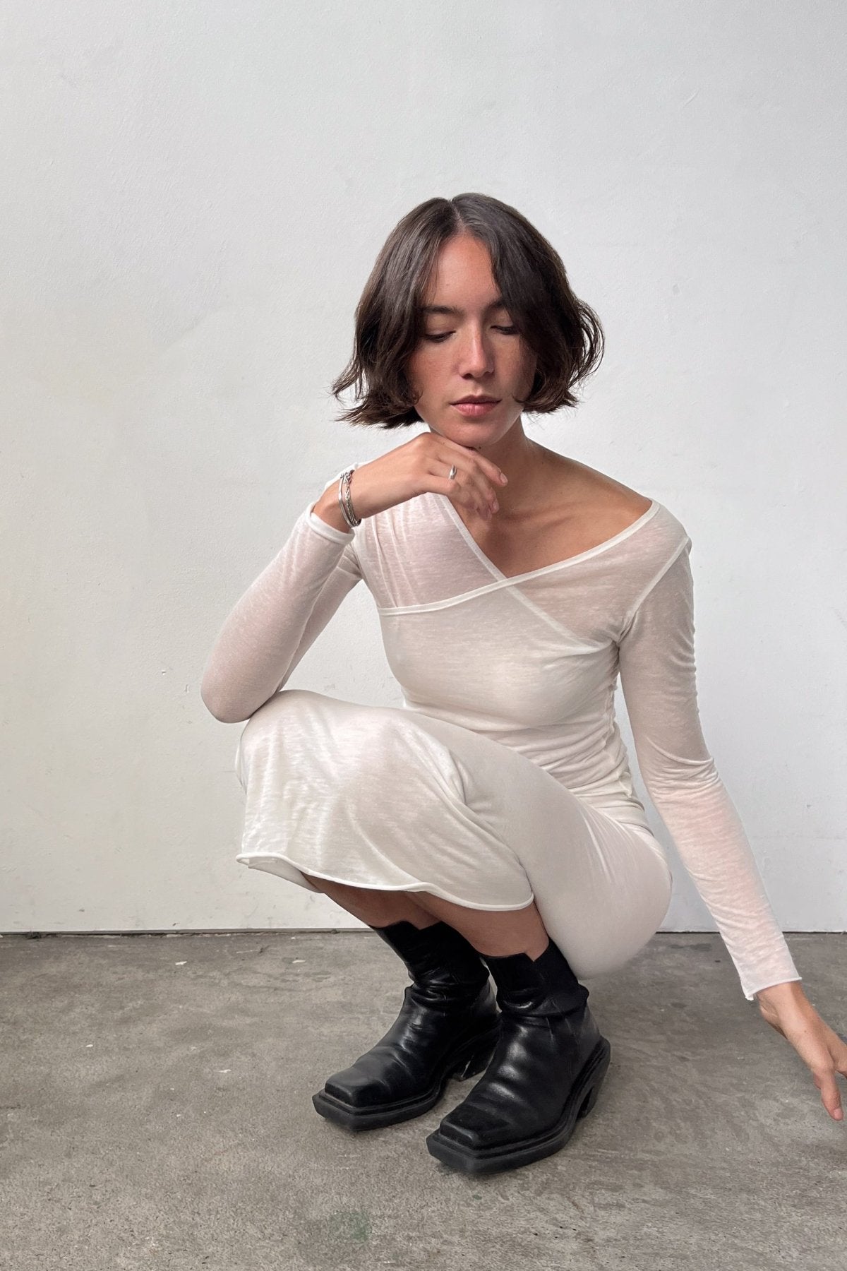 Model kneeling down, wearing the Asymmetric V-Neck Long Sleeve Top in Off White, crafted from a soft blend of recycled wool and modal.
