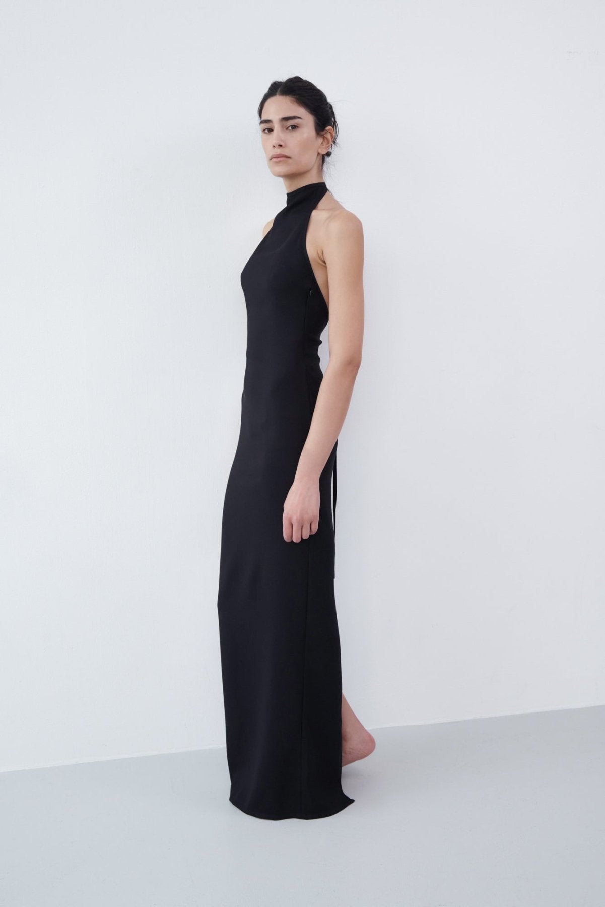 Side profile of a black halter maxi dress with a high neckline and sleek, fitted silhouette, styled barefoot against a minimalist backdrop.