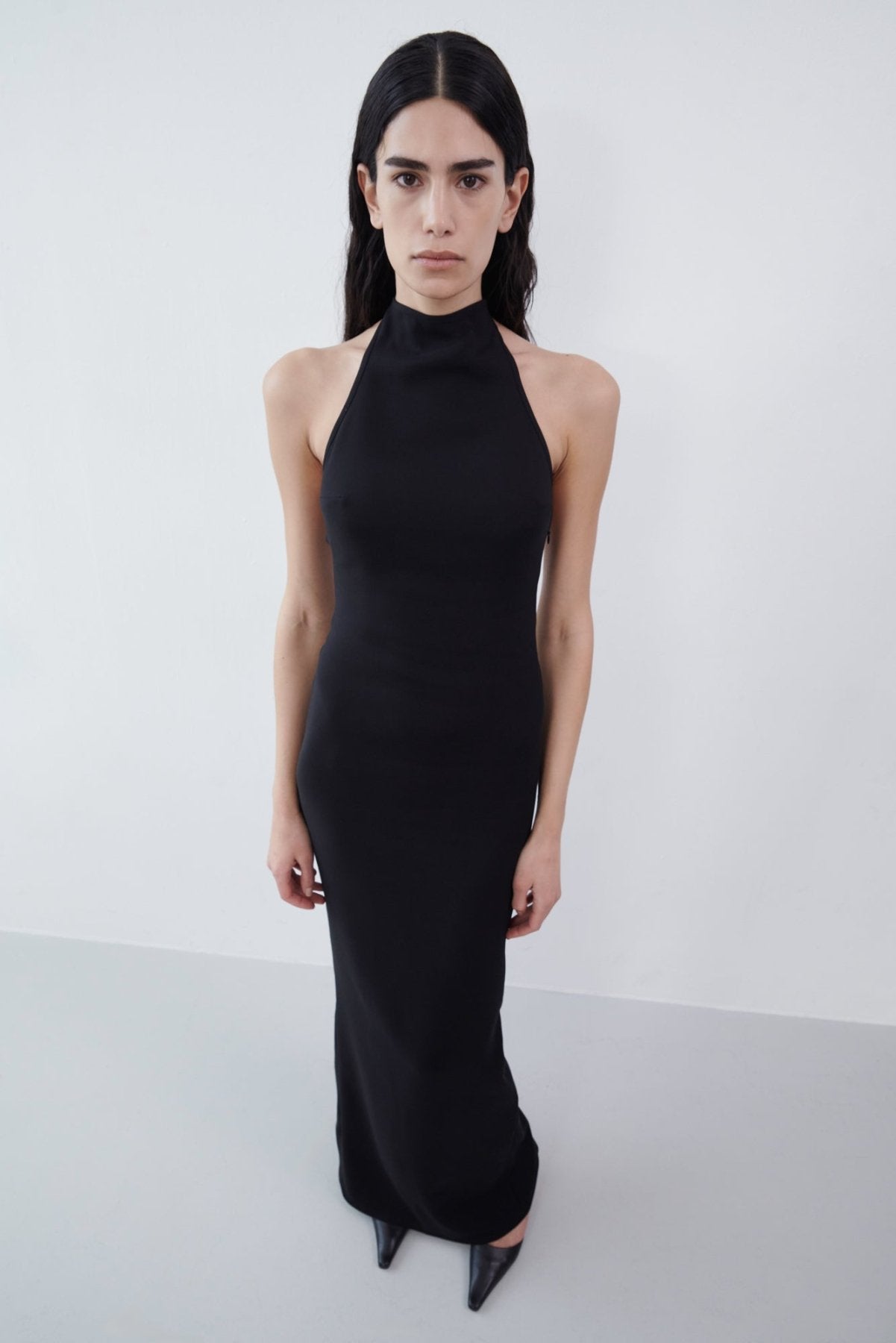 Front view of a black halter maxi dress styled with pointed black heels, featuring a high neckline and a sleek, floor-length silhouette.