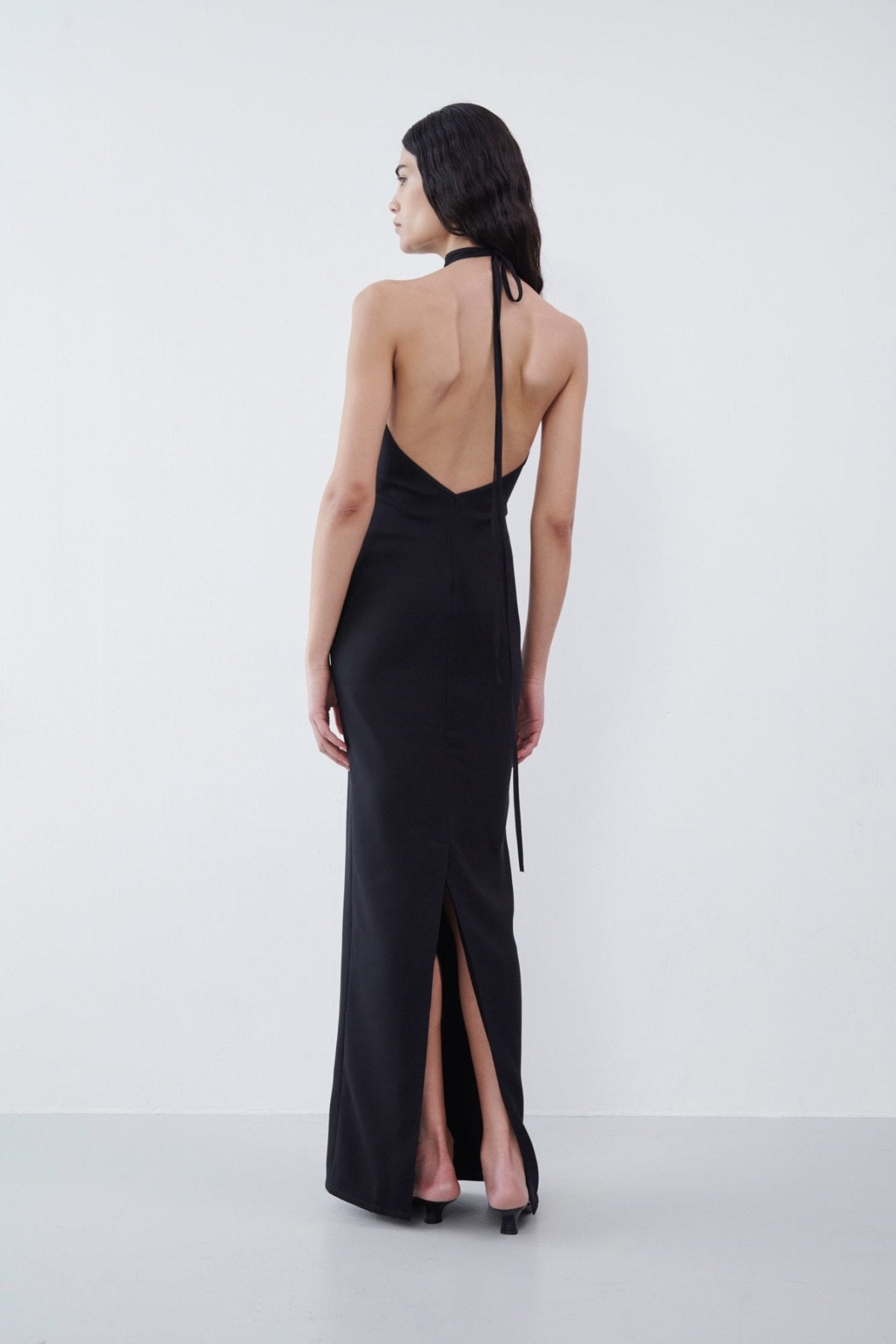 Rear view of a black backless halter maxi dress with a tie detail and a subtle slit at the back hem, styled with black heels.