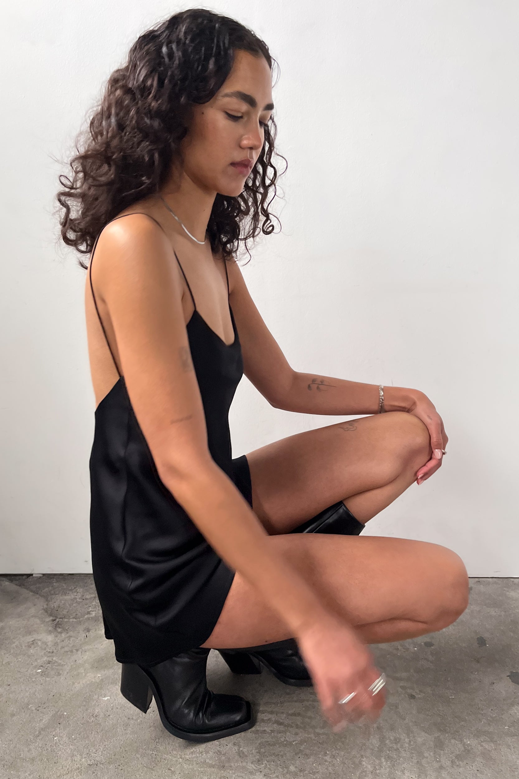 Side profile of the model kneeling down in our Backless Short Slip Dress in black, highlighting its soft and fluid satin material.